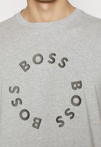 Šedé bavlněné tričko s kulatým černým nápisem "BOSS", uspořádaným do kruhu, s texturovaným perforovaným designem.