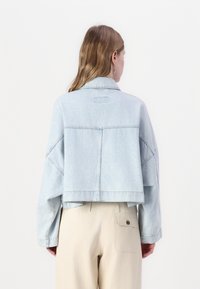 Giacca di denim azzurro chiaro, taglio corto, con colletto ribassato, spalle scese e dettagli di cucitura sottili, abbinata a pantaloni beige.