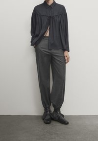 Zwarte overhemd met knoopsluiting en een strik in de taille, gecombineerd met losse grijze broek en donkere schoenen met rimpelaccenten. Stijlvolle stofstructuren.