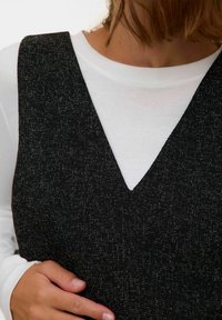 Juodas tekstūruotas suknelė su V formos iškirpte, uždėta ant baltos ilgomis rankovėmis marškinio. Matoma ranka ant liemens prideda gylio.