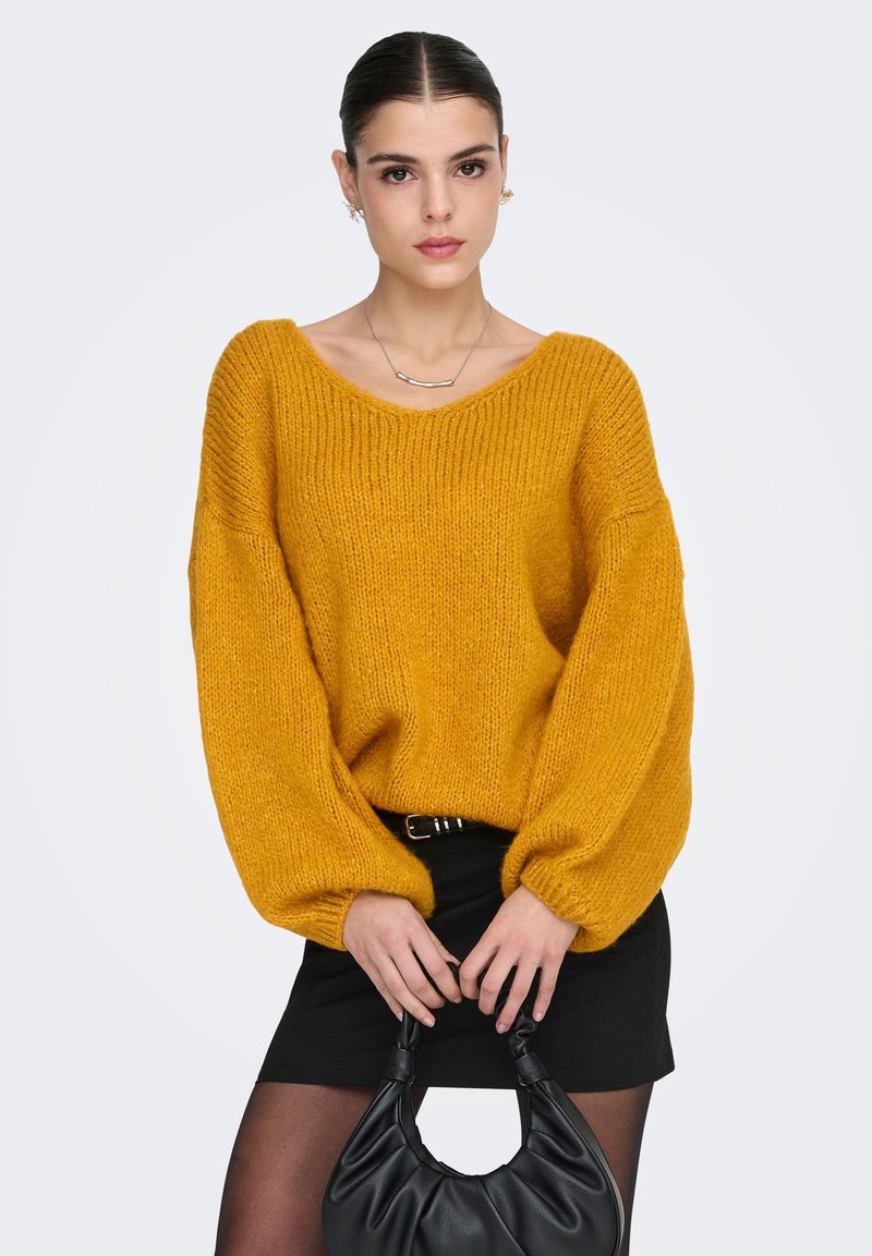 Sennepsgul oversized strikkegenser med bred hals og voluminøse ermer, kombinert med et svart miniskjørt og en svart håndveske.