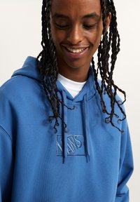 Sudadera azul fabricada en un tejido suave, con un logo bordado de Vans en el pecho. Tiene un bolsillo canguro y capucha con cordón.
