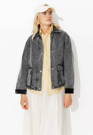 Veste en denim grise avec des poignets noirs, des poches avant et des boutons argentés ; superposée à une chemise à rayures jaunes et une jupe en dentelle blanche.