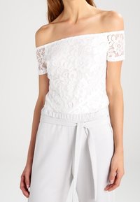 Femme portant un haut en dentelle blanc à épaules dénudées avec un motif floral et un pantalon blanc taille haute avec une ceinture nouée.