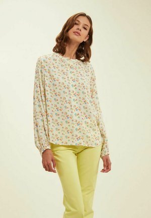 Blouse à motif floral en crème clair, avec encolure ronde, manches longues avec poignets élastiques, et une coupe décontractée. Associée à un pantalon jaune.