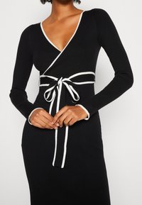 Robe noire côtelée avec manches longues, col en V et bordure blanche. Dotée d'une taille nouée pour un ajustement réglable et une silhouette élégante.