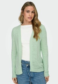 Woman with long blonde hair wearing a light green open-knit cardigan, white top, and blue jeans against a plain background.