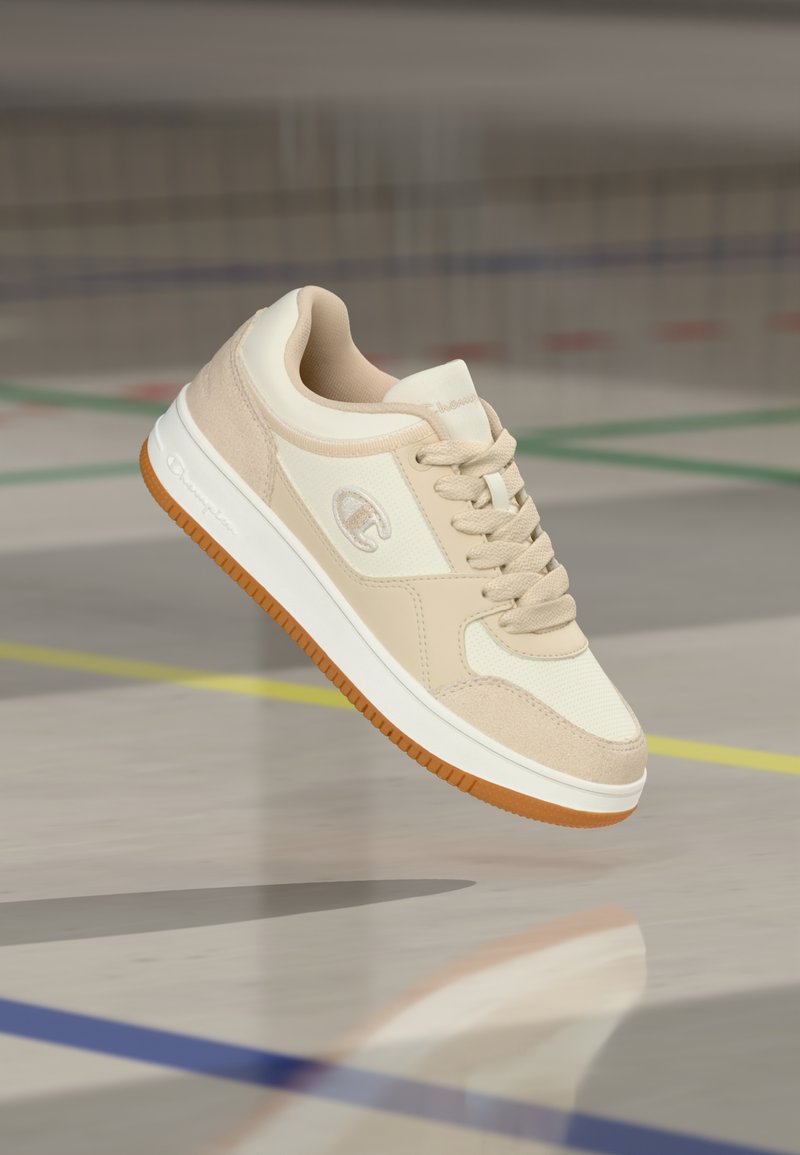 Μπεζ και λευκό αθλητικό παπούτσι με καουτσούκ σόλα, δεμένα κορδόνια, αιωρούμενο πάνω από δάπεδο γυμναστηρίου σημαδεμένο με έγχρωμες γραμμές.