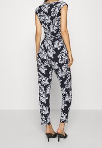 Woman wearing a black and white floral print jumpsuit and black high heel mules, standing against a plain white background.