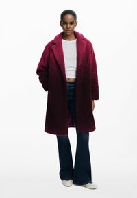 Burgunderfarbener, flauschiger Mantel mit großen Revers, einem Knopfverschluss und zwei Taschen, kombiniert mit hellen, weit geschnittenen Jeans und weißen Schuhen.