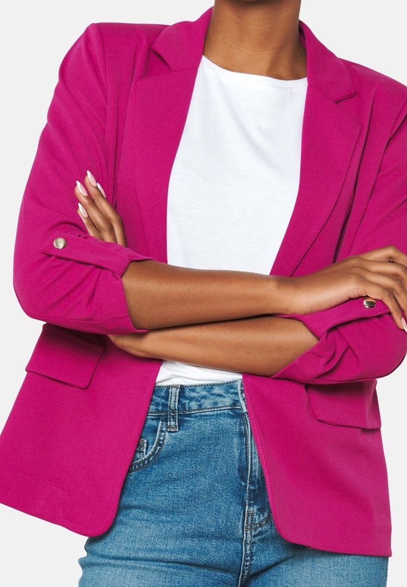 Blazer fucsia con diseño estructurado, cuello con solapa, mangas remangadas y bolsillos frontales, combinado con una camisa blanca lisa y jeans azules.