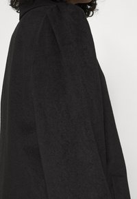 Chaqueta negra de manga larga con tejido texturizado, que presenta un cuello alto y un ligero acolchado en los hombros, con bolsillo lateral visible.