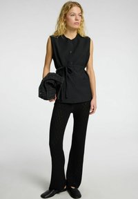 Top negro sin mangas con botones y un cinturón tipo lazo, combinado con pantalones acampanados negros texturizados. La modelo sostiene una chalina negra texturizada.