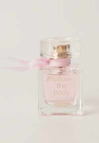 Przezroczysta szklana butelka perfum o kwadratowej podstawie, złotych akcentach i różowej wstążce. Płyn wewnątrz ma delikatny różowy kolor. Etykieta wskazuje "#follow the body."