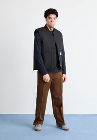 Melnā bezpiedurkņu veste pāri tumši zilai garajai krekla, sakombinēta ar brūniem biksēm. Modelis valkā pelēkas sporta kurpes; vienkārša fona.