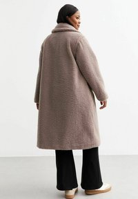 Lungo cappotto beige chiaro realizzato in tessuto felpato, con un ampio colletto e maniche lunghe. Indossato sopra pantaloni neri e calzature chiare.