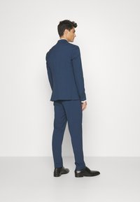 Navy suit with a tailored fit, single vent at the back, and slim trousers. Black shoes complete the outfit, shown from behind against a white background.