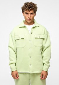 Lichtgroene jas van zachte stof, voorzien van twee voorzakken, drukknoopsluitingen en een relaxte pasvorm. Gedragen over een wit shirt.