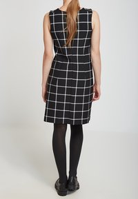 Robe sans manches à carreaux noirs et blancs avec une silhouette ajustée, présentant un ourlet au niveau du genou et accompagnée de collants opaques noirs et de chaussures.