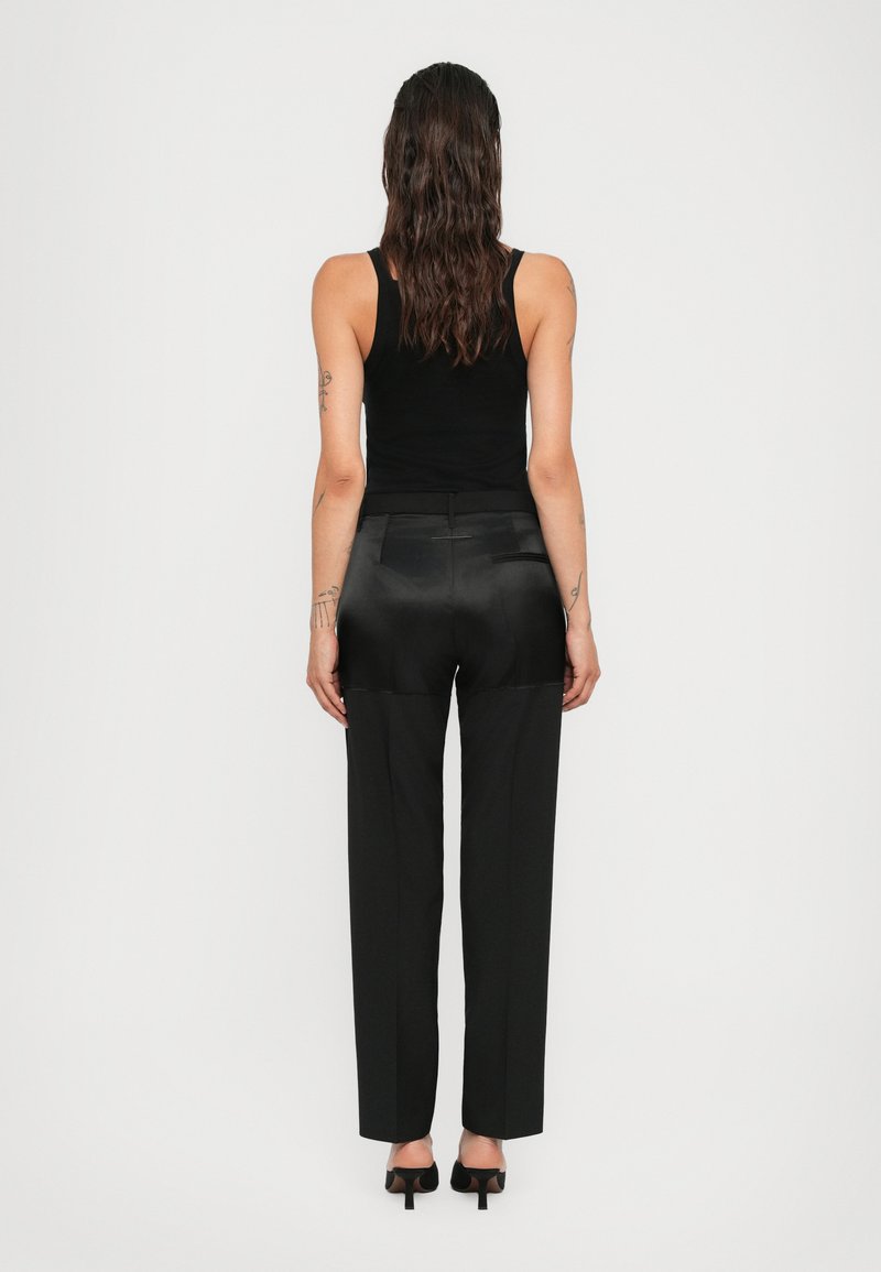 Femme aux longs cheveux bruns portant un haut sans manches noir, un pantalon noir avec garniture en satin et des chaussures à talons noires, se tenant de dos sur un fond uni.
