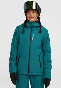 Teal skijas met gestructureerde stof, ritszakken, verstelbare capuchon en zwarte handschoenen. Model draagt skibril en heeft een ingevlochten haar.