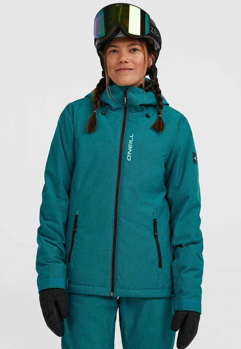 Teal skijas met gestructureerde stof, ritszakken, verstelbare capuchon en zwarte handschoenen. Model draagt skibril en heeft een ingevlochten haar.