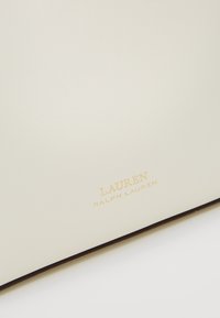 Krémová kožená textura se zlatě embosovaným logem "LAUREN RALPH LAUREN" na hladkém povrchu, pravděpodobně kabelka nebo doplněk.