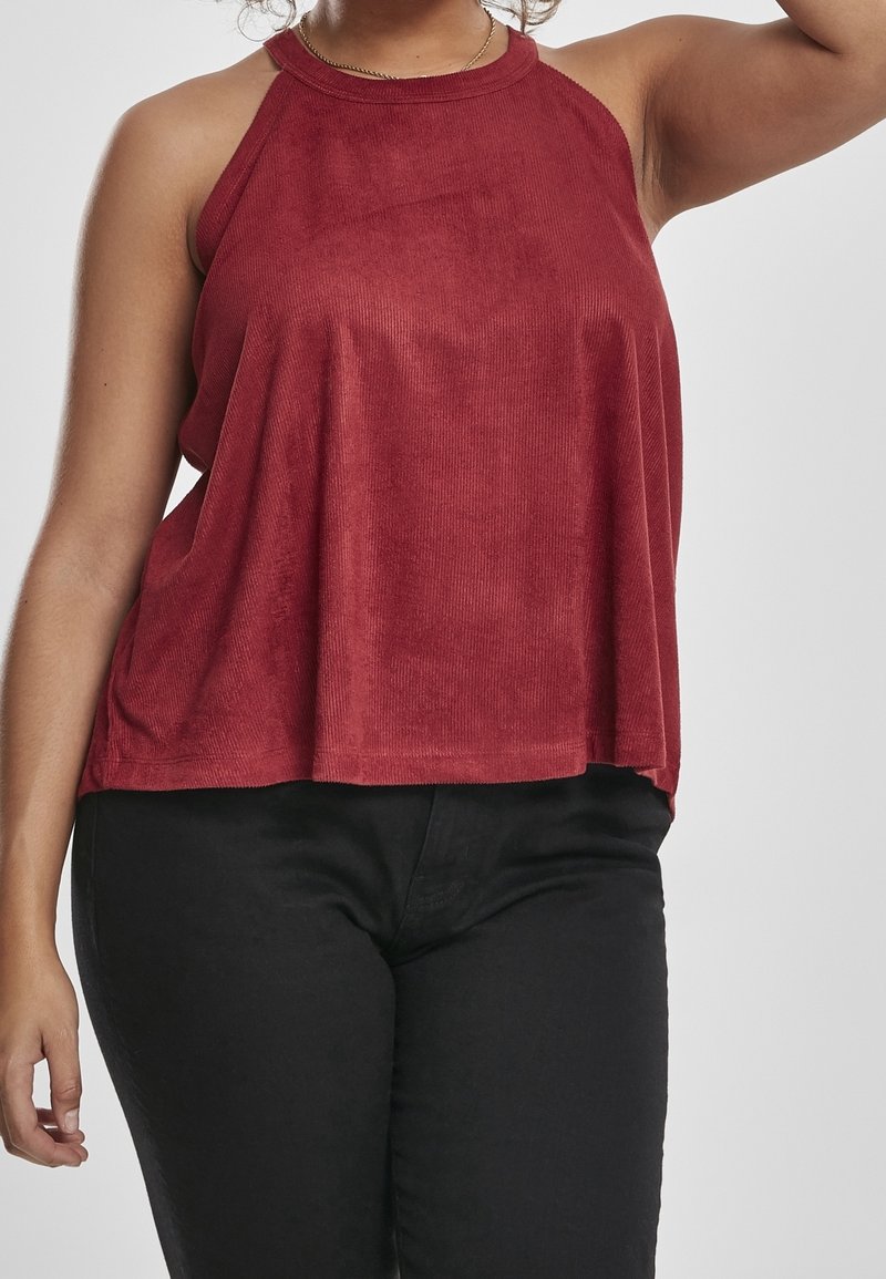 Femme portant un haut rouge sans manches et un pantalon noir, avec un bras levé contre un fond clair uni.