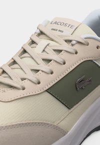 Baskets Lacoste beige et vert avec lacets beige, panneaux en mesh et cuir, arborant un logo crocodile métallique sur le côté.