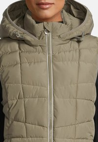 Gilet rembourré beige avec capuche, présentant une fermeture éclair verticale et une texture matelassée. Comprend des détails à cordon de serrage sur la capuche.