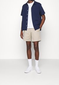 Kortärmad marinblå skjorta med vita knappar, tillsammans med beige shorts. Vita sneakers och ankelsockor kompletterar outfiten, mot en neutral bakgrund.