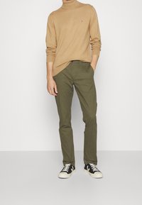 Beżowy sweter z golfem, oliwkowe spodnie i czarne sneakersy z białymi akcentami. Spodnie mają kieszeń z przodu; sweter ma logo.