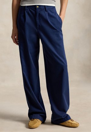 Personne portant un pantalon bleu marine ample et des mocassins couleur beige, debout avec une main dans la poche sur un fond neutre.