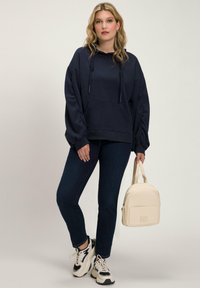 Marineblauwe oversized hoodie met een voorzak, trekkoorden en geribde manchetten, gedragen met donkerblauwe jeans en een crèmekleurige rugzak. Witte sneakers.