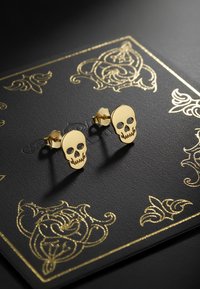 Orecchini a forma di teschio oro con dettagli neri sugli occhi, su uno sfondo nero texturizzato con motivi floreali dorati.