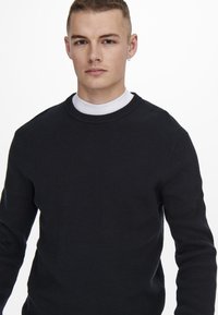 Sweater noir à côtes avec un col rond, présentant un motif de tricot texturé. Porté par-dessus un haut à col montant blanc, affiché sur un fond neutre.