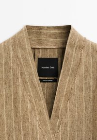 Chemise en lin brun avec un décolleté en V, présentant une finition texturée et une étiquette de marque noire à l'intérieur du col. Détails de couture visibles.