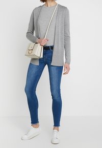 Ljust grå cardigan med öppen front, i kombination med en grå t-shirt, blå slim-fit jeans och vita sneakers. En liten beige crossbody-väska bärs.