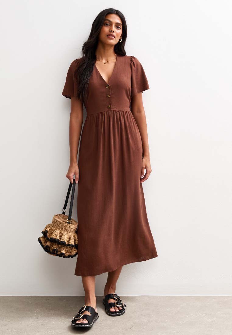 Vestido midi marrón con mangas cortas abullonadas, cierre de botones en la parte delantera y cintura fruncida. Combinado con un bolso de paja y sandalias negras. Textura suave.