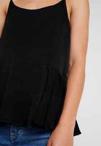 Top negro sin mangas con un acabado texturizado, que presenta un dobladillo de peplum y tirantes finos, combinado con unos jeans de mezclilla azules.