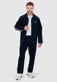 Marineblauwe ribfluwelen trainingsoutfit met een ritsjack met witte strepen en bijpassende broeken, beide met logo-accenten en geribde manchetten.