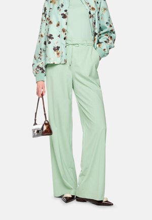 Woman wearing light green wide-leg pants and floral jacket, holding a small brown and white handbag in one hand.