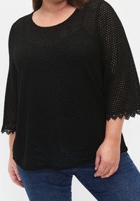 Femme portant un haut en maille crochet noire avec des bords festonnés sur les manches trois-quarts et un jean bleu foncé, sur un fond uni.