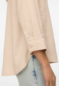 Chemise beige à manches longues avec une coupe décontractée, dotée d'un poignet boutonné et d'un ourlet arrondi. Associée à un jean léger bleu déchiré.