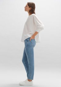 Chemisier blanc léger à manches longues avec un ourlet arrondi, assorti à un jean skinny bleu clair et des baskets blanches.