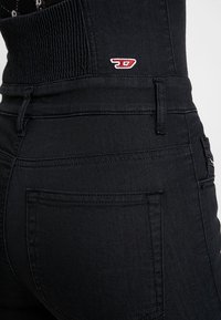 Gros plan sur un pantalon noir taille haute avec poche arrière cousue et petit logo rouge et blanc près de la ceinture.