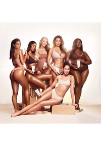 Six diverse women in neutral-toned underwear hold skincare tubes, posing confidently with soft lighting and wooden boxes on a plain background.