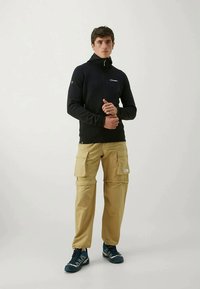 Casaco de fleece preto com fecho, mangas longas. Calças de sarja castanhas com vários bolsos. Ténis desportivos azuis. De pé contra um fundo simples.