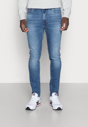 Man met lichtblauwe slim-fit jeans en witte New Balance sneakers, staand tegen een effen achtergrond.