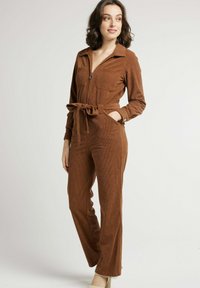 Combinaison en velours côtelé marron avec une fermeture zippée à l'avant, des manches longues, deux poches poitrine et une ceinture à la taille. Présente une silhouette ajustée et des jambes évasées.